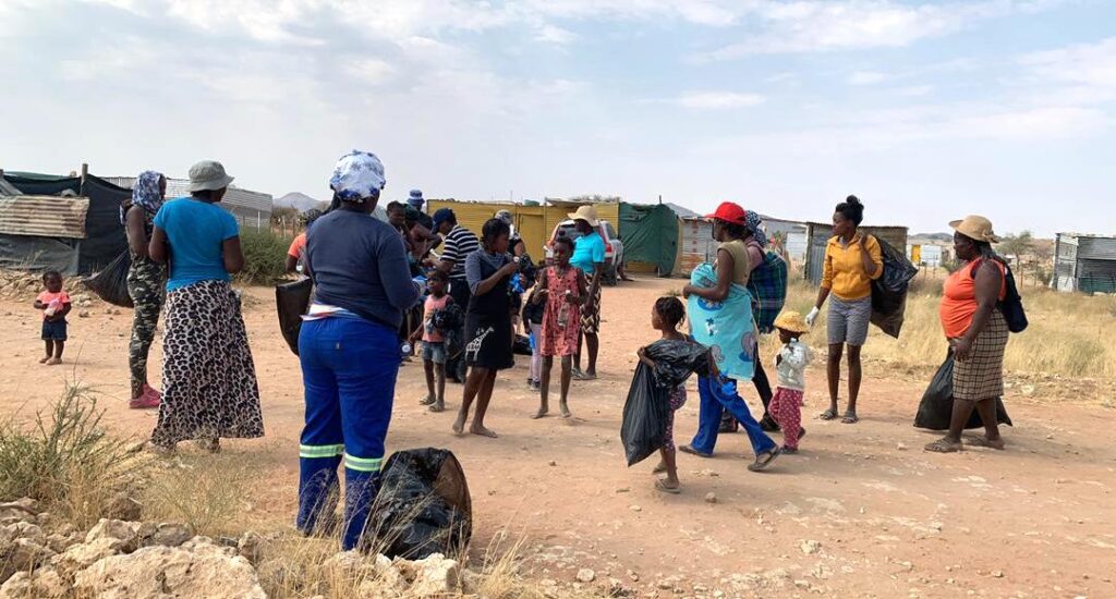 A group of people carrying black sacks with several shacks in the background.