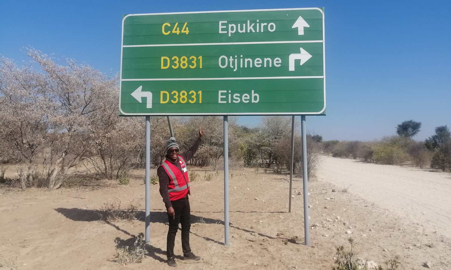A man in a red hi-vis jacket points at a green road sign.
